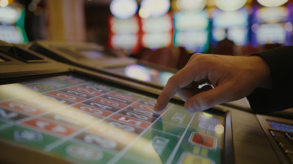 Hand hovering over video poker touchscreen mid-decision on a blurred casino floor